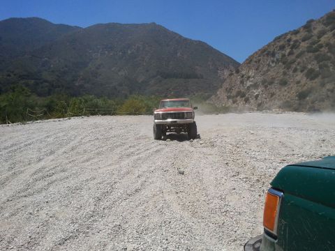 1978 Ford Bronco - A red pickup truck kicks up dust on a gravel road, with another vehicle visible in the foreground.