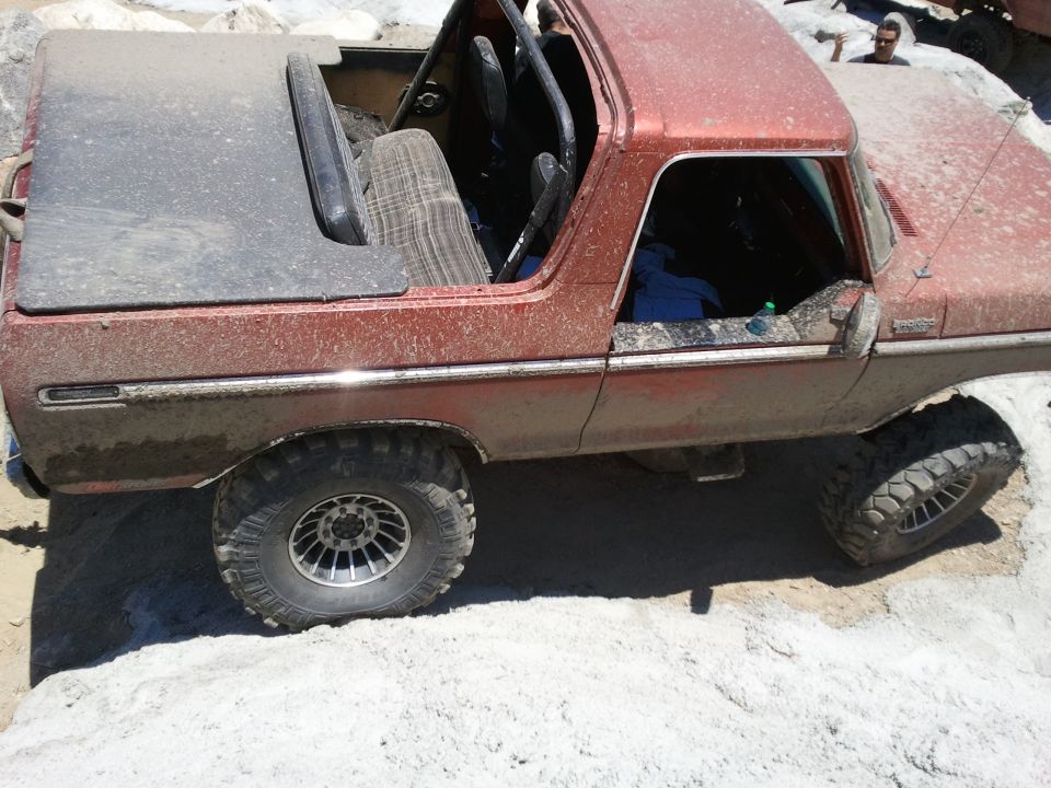 Red 1978 Ford Bronco with a soft top and roll cage, covered in dirt, on a rocky terrain
