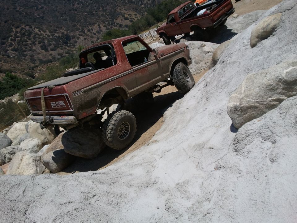 Red 1978 Ford Bronco climbing rocks, with a red pickup truck behind it