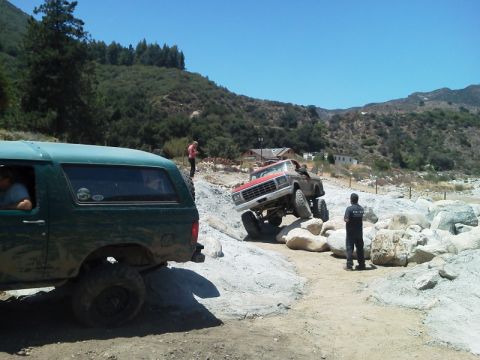 1978 Ford Bronco - Two trucks are shown off-roading on a sunny day, with one truck climbing over large rocks.