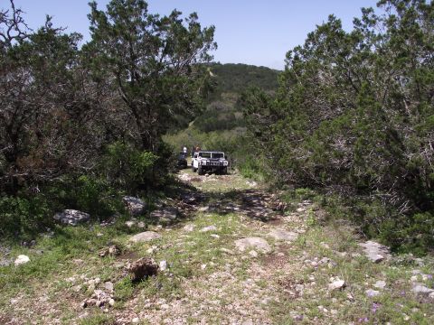 1989 Ford Bronco - Off-road vehicles navigate a challenging rocky trail through a wooded, hilly landscape.