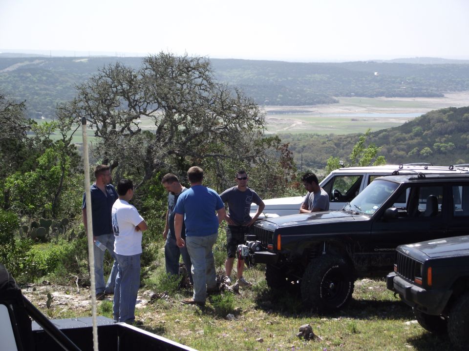 Several Jeep Cherokees parked on a grassy hill with people standing around them; a valley is visible in the background