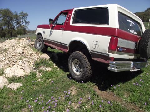 1989 Ford Bronco - A two-tone red and white Ford Bronco navigates a rocky, grassy incline under a clear blue sky.