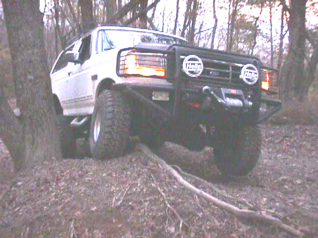 White Ford Bronco flexed up over a log obstacle on a trail, round driving lights on the brush guard