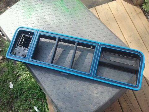 1979 Ford Bronco - This is a close-up of a vehicle's grille assembly, blue trim and a vent. The part is resting on a textured surface outdoors.