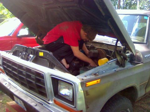 1979 Ford Bronco - A person works on the engine of a brown Ford Bronco, with a red truck visible in the background.