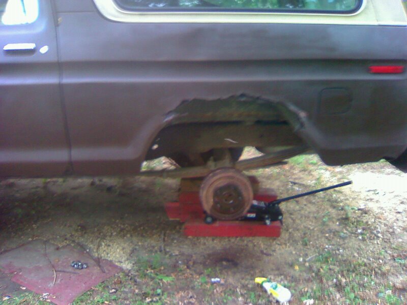 1979 Ford Bronco rear wheel well showing significant rust damage, supported by a jack and wooden blocks