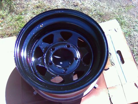 1979 Ford Bronco - A freshly painted black steel wheel sits on a cardboard surface outdoors.