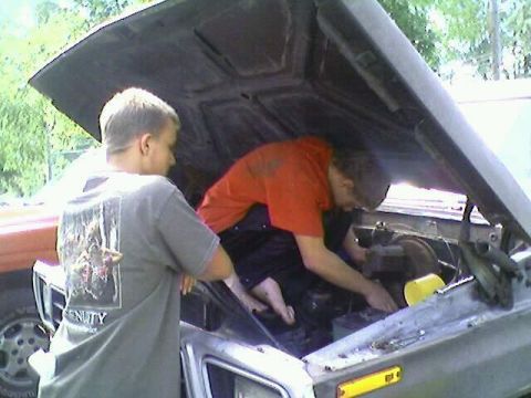 1979 Ford Bronco - Two individuals are actively working on the engine of a vehicle, with the hood open.
