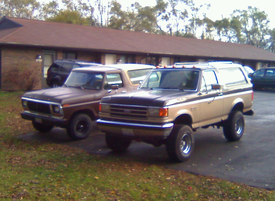 Two Ford Broncos parked side-by-side. The front Bronco is brown. The rear Bronco is tan with a black stripe and lifted suspension