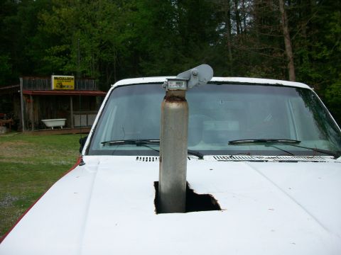 1985 Ford Bronco - A custom exhaust stack protrudes through the hood of a white truck, with a rustic building in the background.