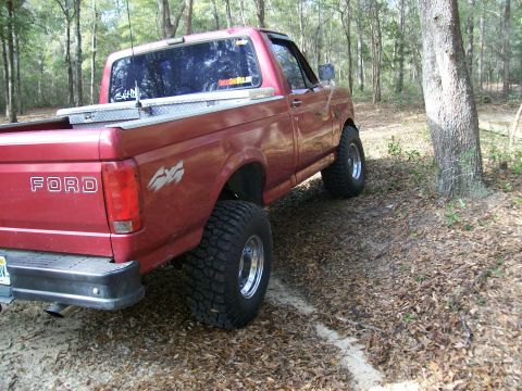 1993 Ford F-150 - Rear view of a lifted red Ford pickup truck with off-road tires and a toolbox in the bed, parked in a wooded area.