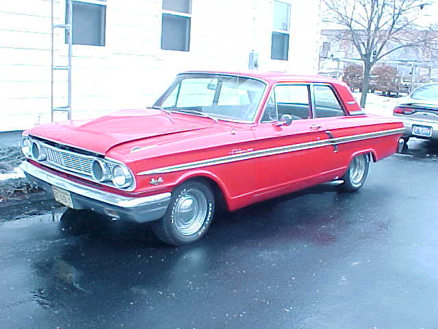 Red 1964 Ford Falcon sedan parked on a wet asphalt surface, viewed from the front three-quarter angle