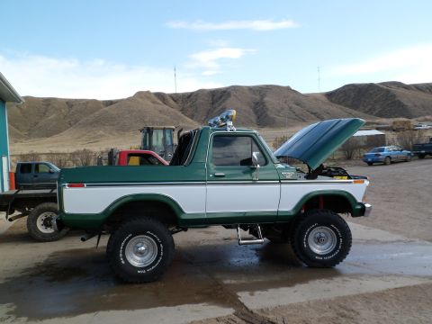 1979 Ford Bronco - A lifted green and white Ford Bronco sits outside with its hood open, parked near other vehicles and machinery.