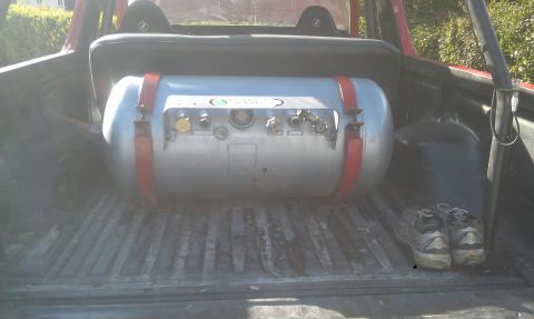 1978 Ford Bronco - A large propane tank is secured with red straps in the bed of a red vehicle, next to a pair of shoes.