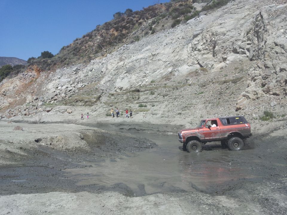 Orange 1978 Ford Bronco driving through a muddy area with a rocky hill in the background
