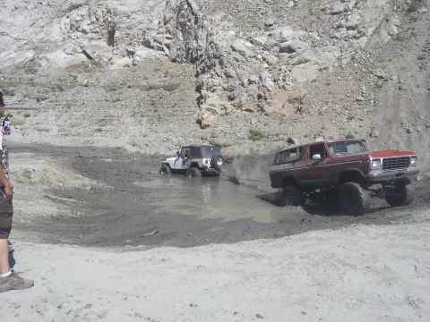 1978 Ford Bronco - Two off-road vehicles navigate a muddy terrain with rocky hills in the background.