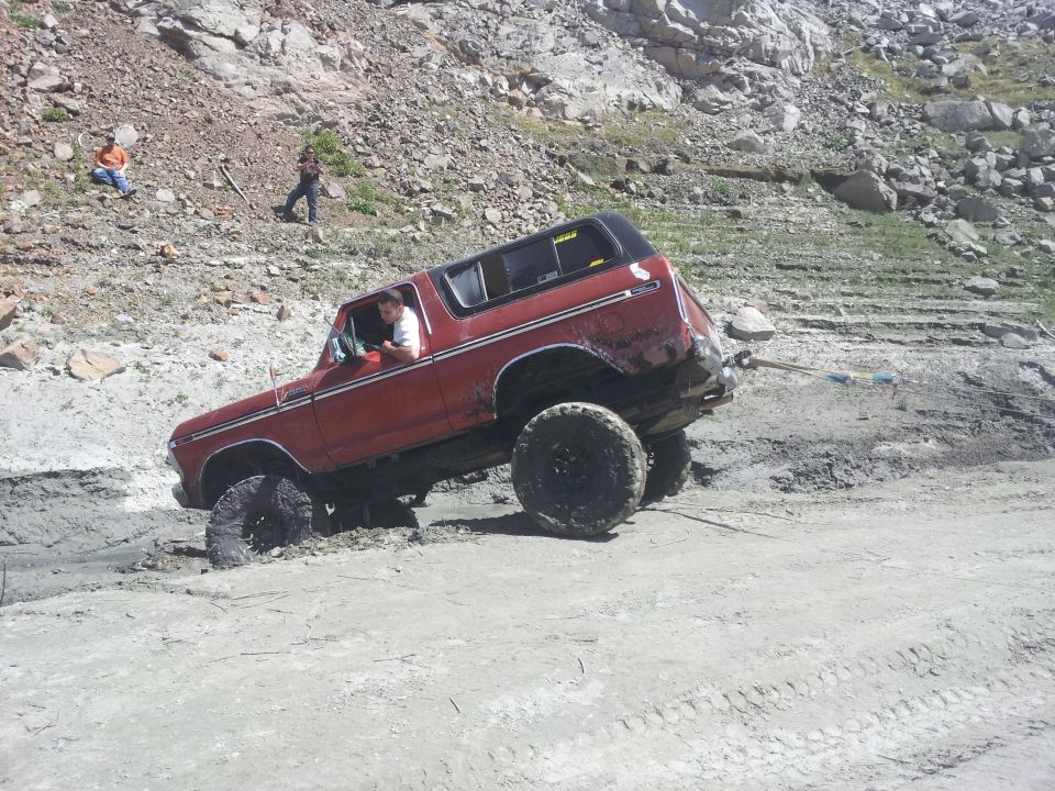 Red 1978 Ford Bronco with large tires stuck in mud on a rocky incline