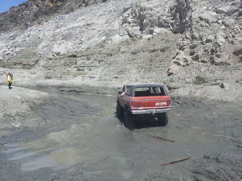 Red 1978 Ford Bronco driving through a muddy area with a rocky hill in the background. A person in a yellow jacket stands on the left