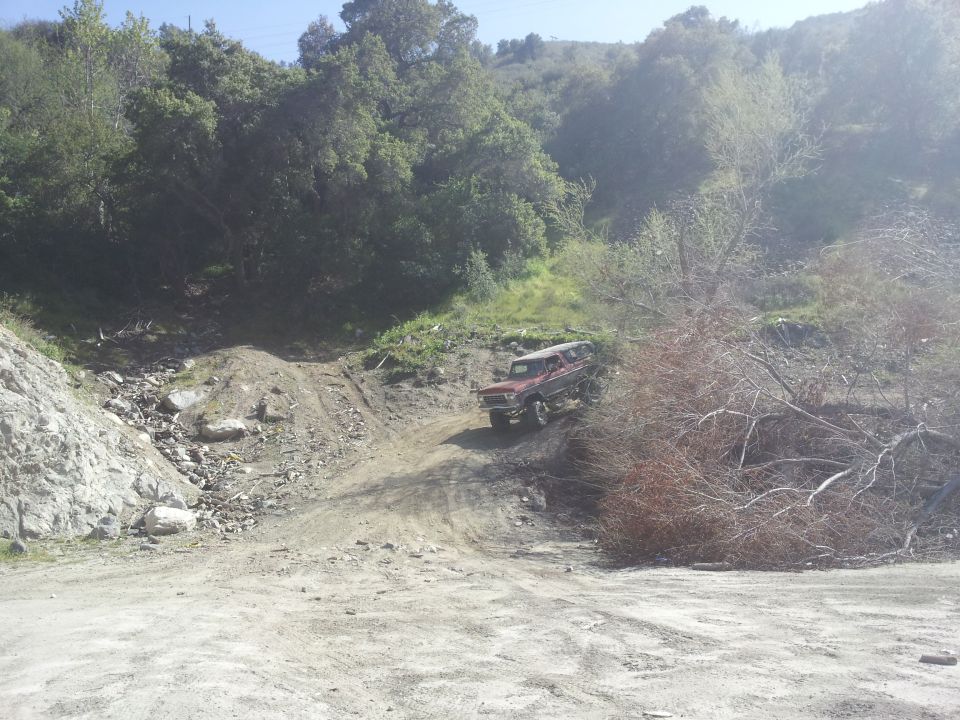 Red 1978 Ford Bronco driving up a dirt hill, front 3/4 view, some brush on the right