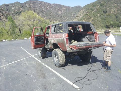 1978 Ford Bronco - A man works on the open tailgate of a red 1978 Ford Bronco parked in a lot with mountains in the background.