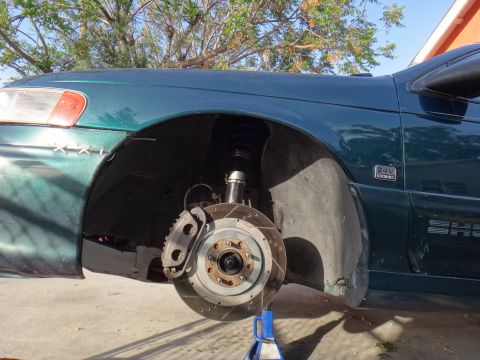 1995 Ford Taurus - The front wheel assembly of a green car, coilovers and slotted brake rotors, is visible with the fender removed.