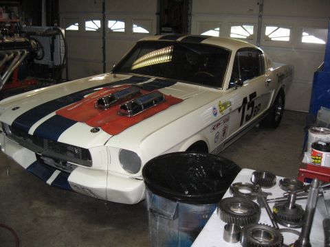 1994 Ford Bronco - White 1965 Ford Mustang race car with blue stripes and dual carburetors is parked in a garage with various parts on a table nearby.