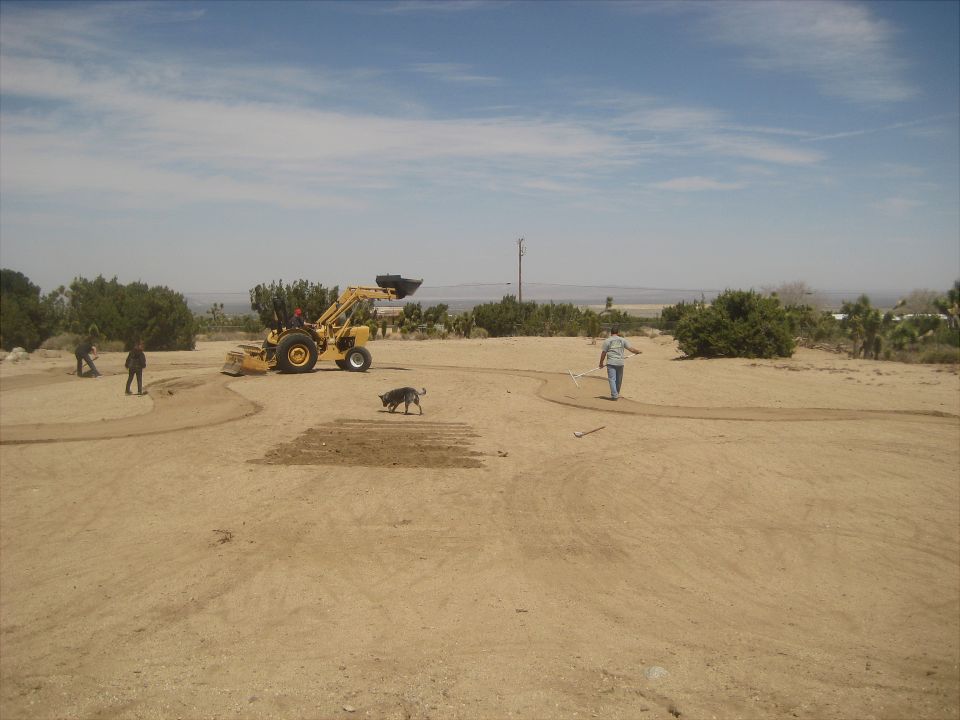 Yellow tractor with front loader, two people, and a dog in a dry, dusty field under a partly cloudy sky