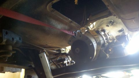 1978 Ford Bronco - The transmission of a vehicle is visible from below, held up by a red strap during what appears to be a repair.