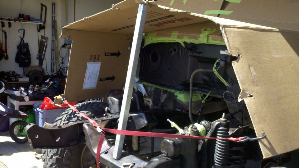 Vehicle chassis with engine bay covered by cardboard, undergoing work. Red straps secure parts
