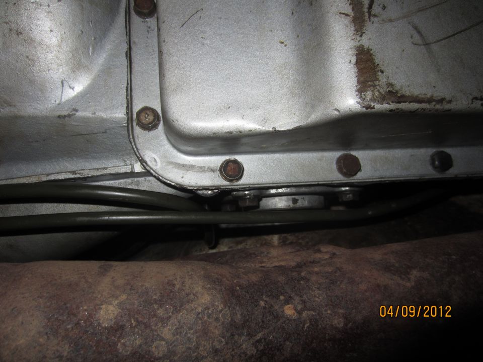 Close-up of a transmission pan with bolts and brake lines, above a rusty exhaust pipe. Date stamp reads 04/09/2012