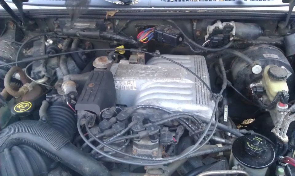 Engine bay with a silver intake manifold, black hoses, and various wires. A yellow oil cap is visible