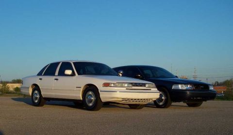 1998 Ford Crown Victoria - Two Ford Crown Victorias, one white and one black, are parked outdoors on a paved surface with a clear blue sky above.