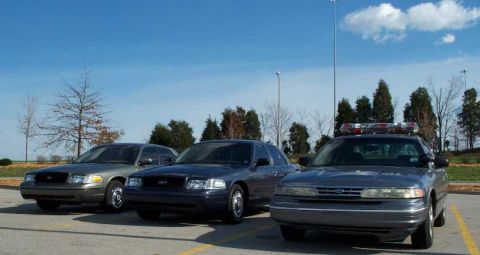 1998 Ford Crown Victoria - Three Ford Crown Victoria police sedans are parked in a lot under a blue sky with scattered clouds.