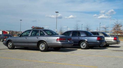 1998 Ford Crown Victoria - Several police sedans are parked in a lot under a cloudy sky, with one vehicle equipped with a light bar.