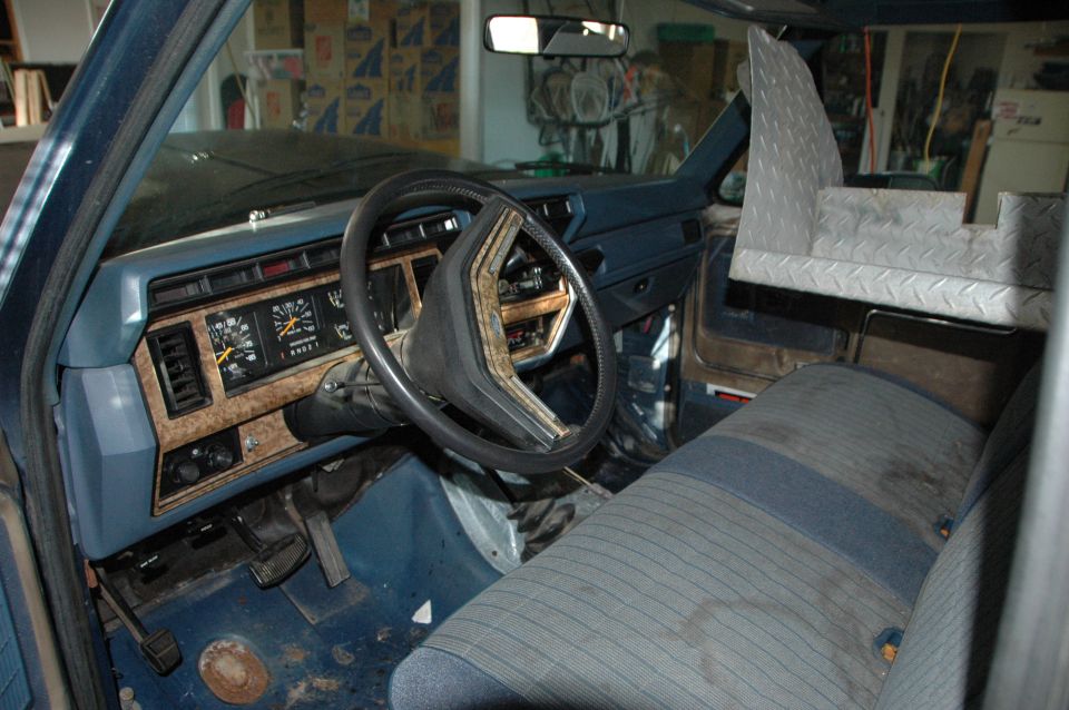 Interior shot of a blue truck or SUV, showing the dashboard, steering wheel, and seats. There's a custom metal storage unit behind the passenger seat