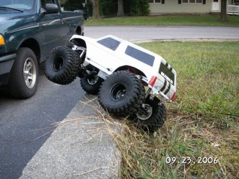 1986 Ford Bronco - RC monster truck attempts to climb a curb, with a real pickup truck nearby.
