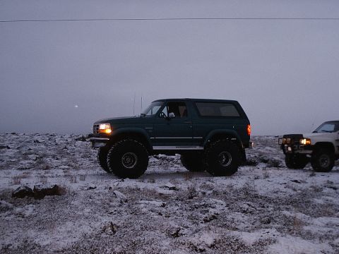 1996 Ford Bronco - Two modified SUVs are parked on a snow-dusted, rocky terrain under an overcast sky.