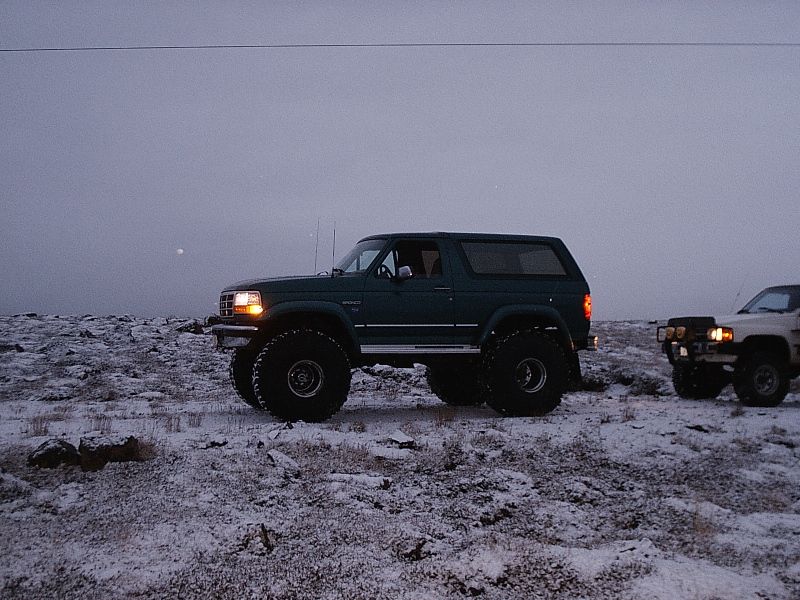 Dark green lifted two-door Ford Bronco SUV, side view, headlights on in snowy field at dusk.