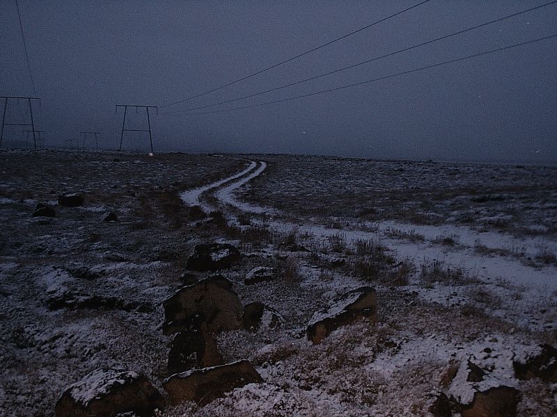 Snow-dusted dirt track winding through barren terrain at dusk with power lines