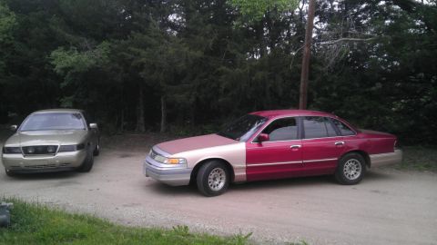 2000 Mercury Grand Marquis - Two sedans, one a red and tan 2000 Mercury Grand Marquis, are parked on a dirt road with trees in the background.