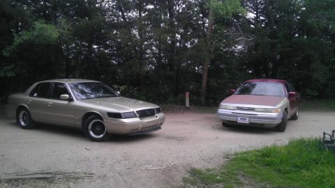 2000 Mercury Grand Marquis - Two sedans, one gold and one maroon/silver, are parked on a dirt road.