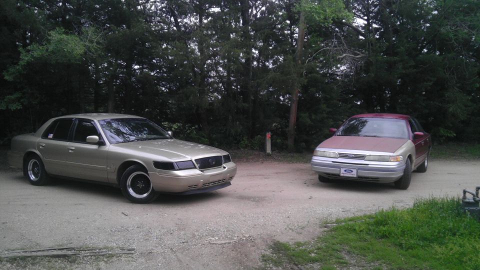 Gold sedan and maroon/silver sedan parked on dirt road with trees in background