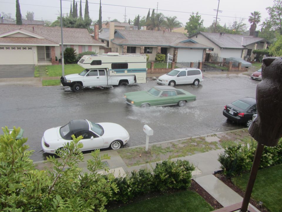 Flooded street with a white convertible, a green sedan, a pickup truck with a camper, and a white minivan driving through the water