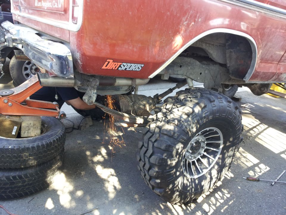 Mechanic sparks flying while cutting leaf spring on red Ford Bronco with large off-road tires
