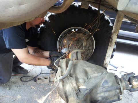 1978 Ford Bronco - A mechanic grinds on the rear axle of a vehicle, with sparks flying and a large tire in view.