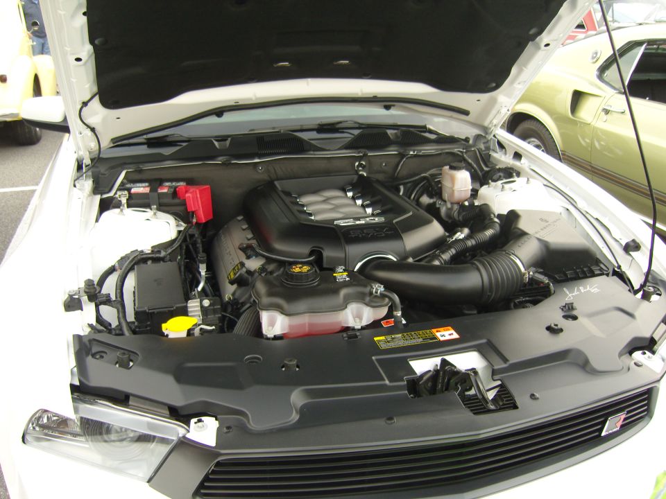 2007 Ford Mustang engine bay with 5.0 badge visible on engine cover. Another classic car is parked to the right