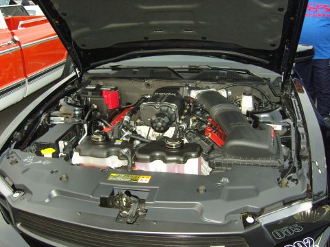 2007 Ford Mustang - The engine bay of a black car, a prominent supercharger and other engine components, with a red and white vehicle partially visible.