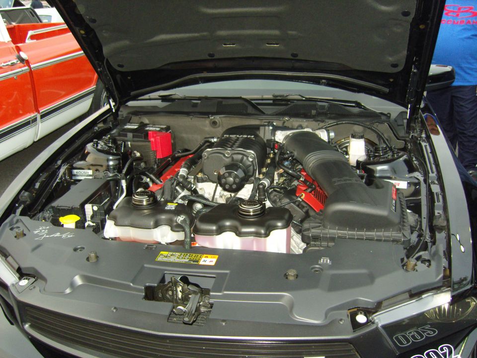 Engine bay of a black car with a supercharger. Another car, red and white, is visible to the left