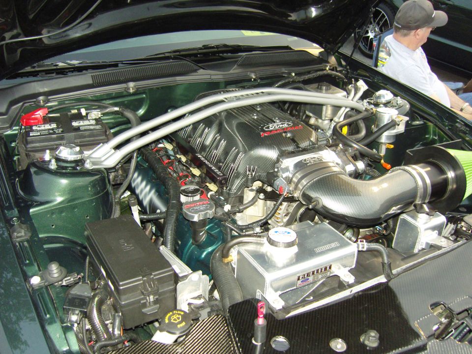 Engine bay of a Ford Mustang with a supercharger and custom intake. A person is visible in the background